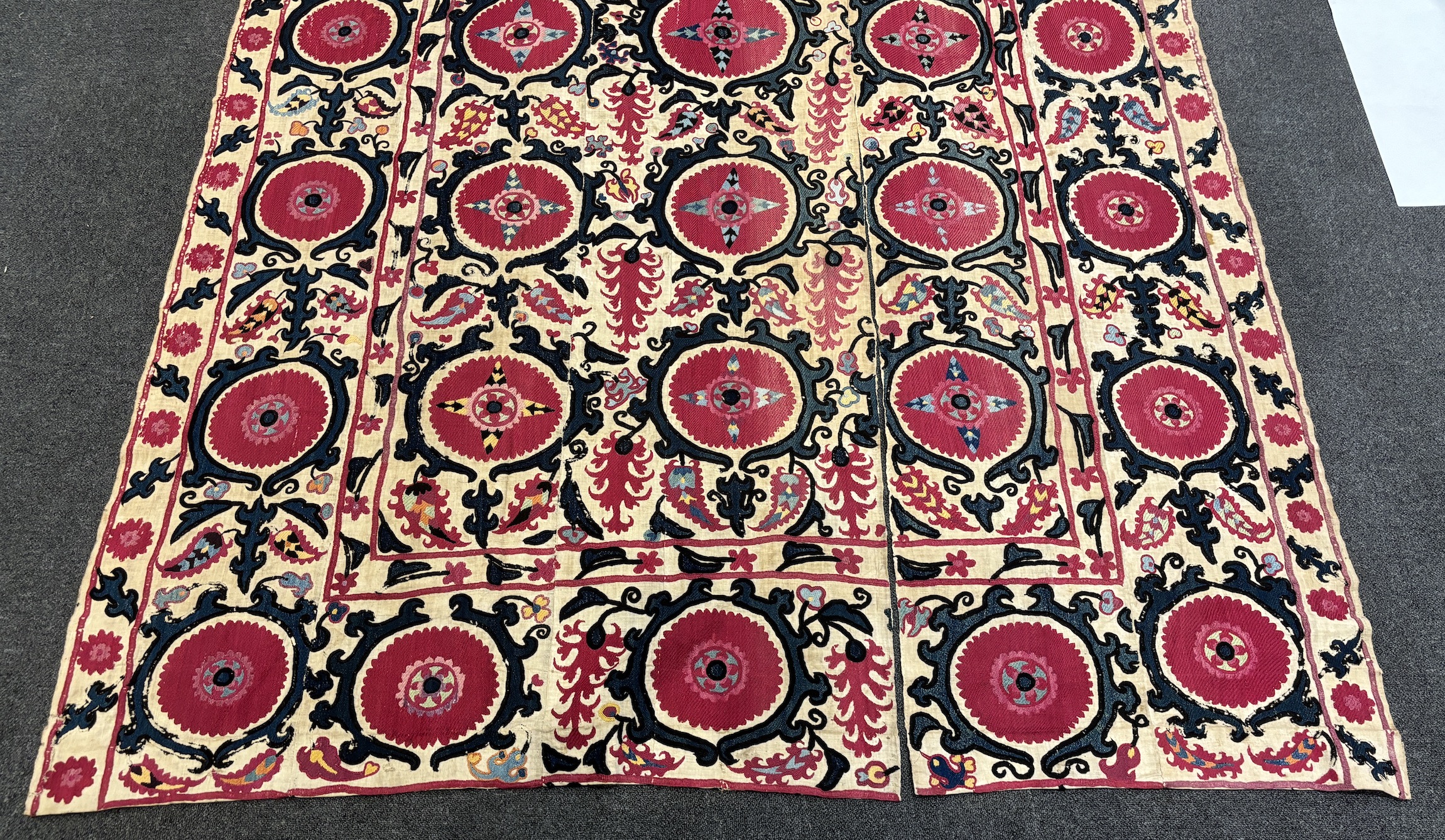 An 19th century Uzbek Suzani embroidered wall hanging, in two pieces, the main hanging sewn together as four panels, together with a fifth matching border, being detached, embroidered with large bold floral motifs, in ri
